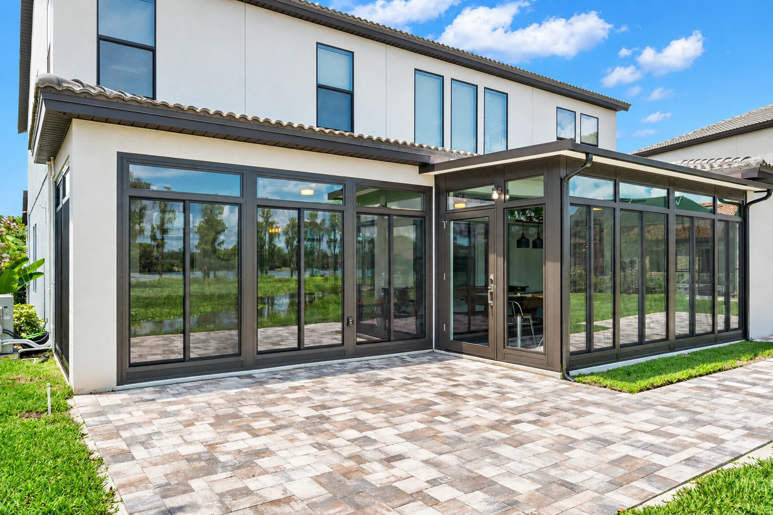 Custom sunroom with dark-framed floor-to-ceiling windows extending along a modern white house beside a paved patio.