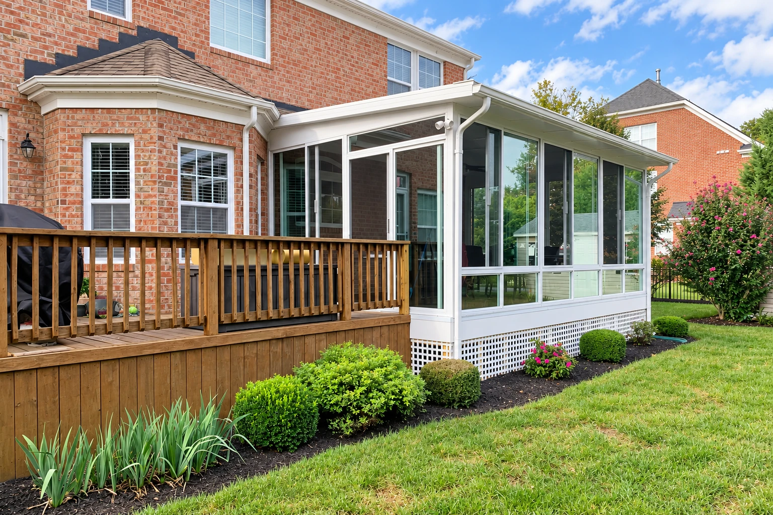 A white-framed sunroom with tall windows is attached to a brick house beside a raised wooden deck and landscaped garden.
