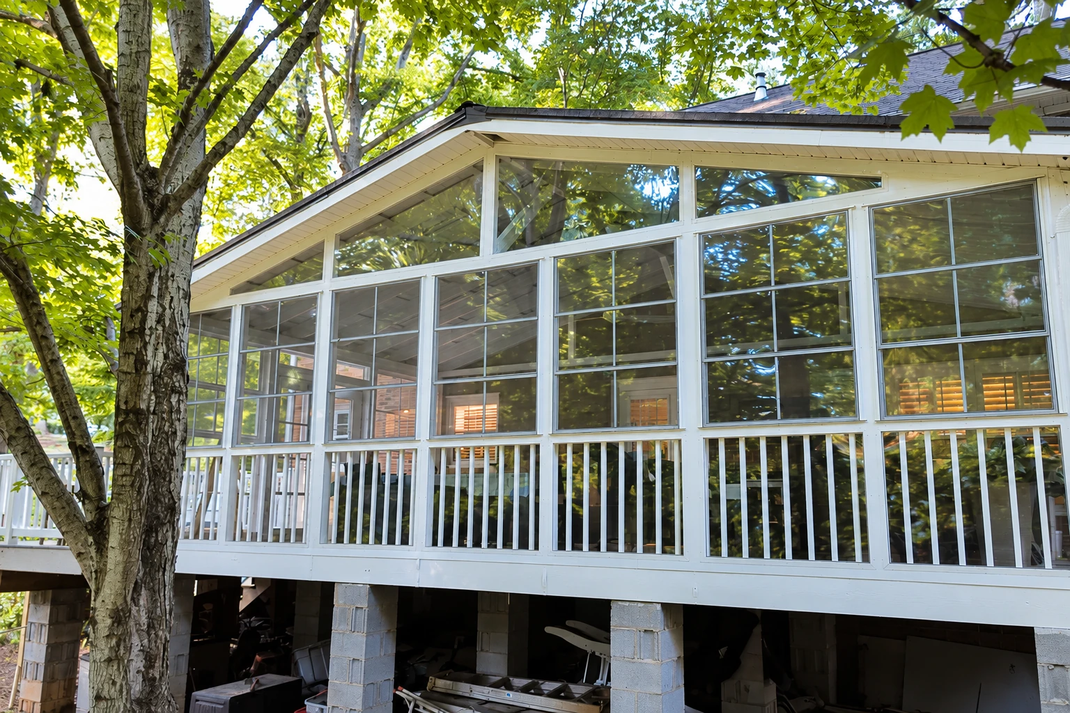 A raised deck enclosed with white-framed windows beneath a peaked roof, surrounded by mature trees.