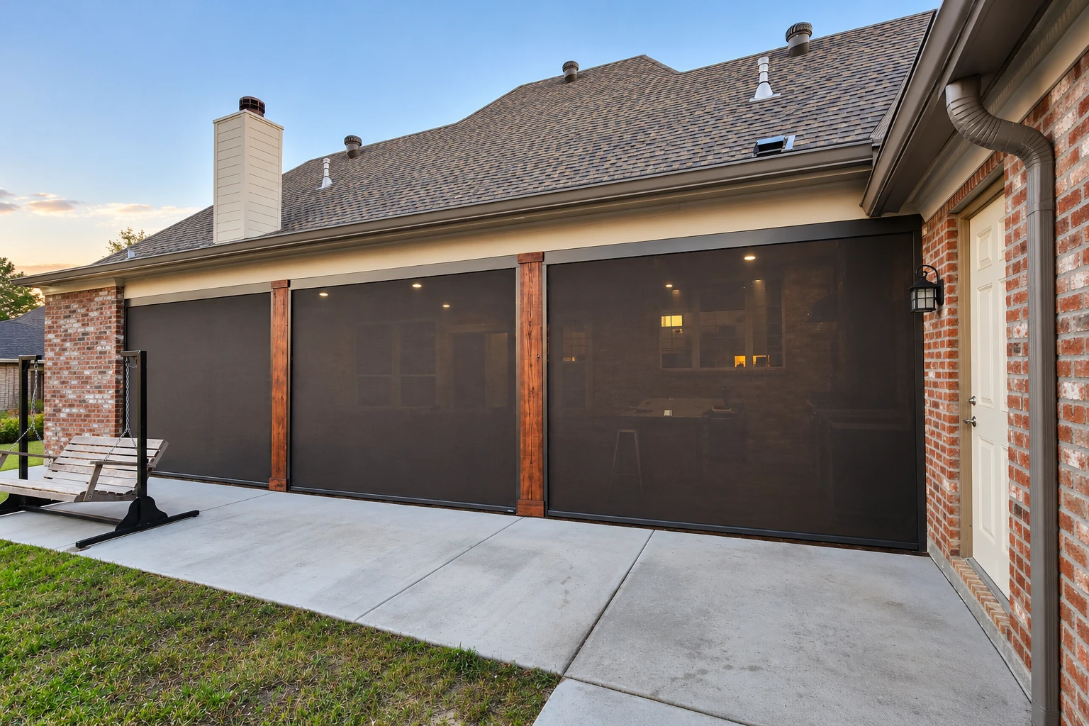Covered patio enclosed with dark mesh screens and wood posts beside a brick house, with a concrete floor and porch swing.