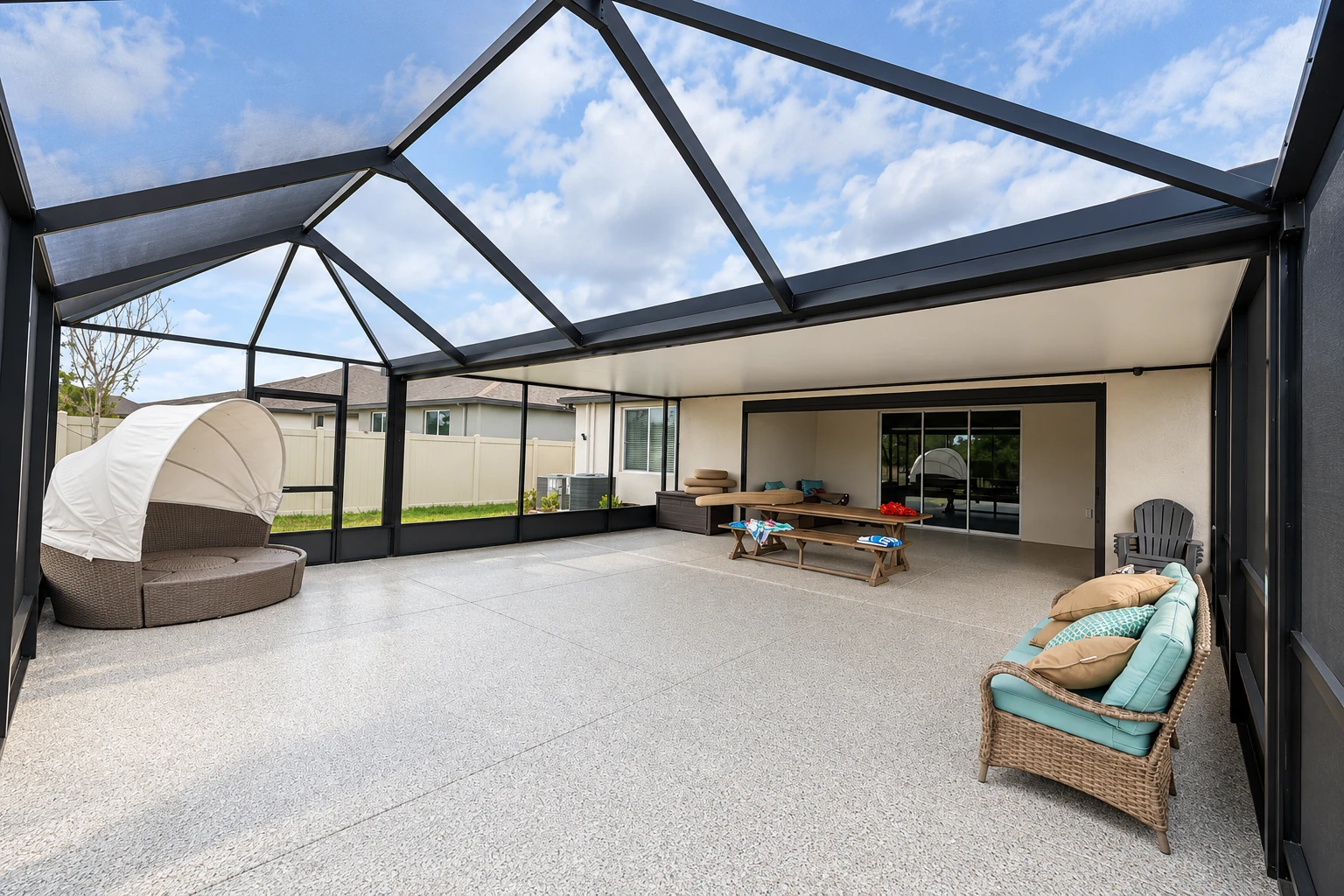 Screened patio enclosure with a dark frame and clear roof, furnished with wicker seating and a picnic table.
