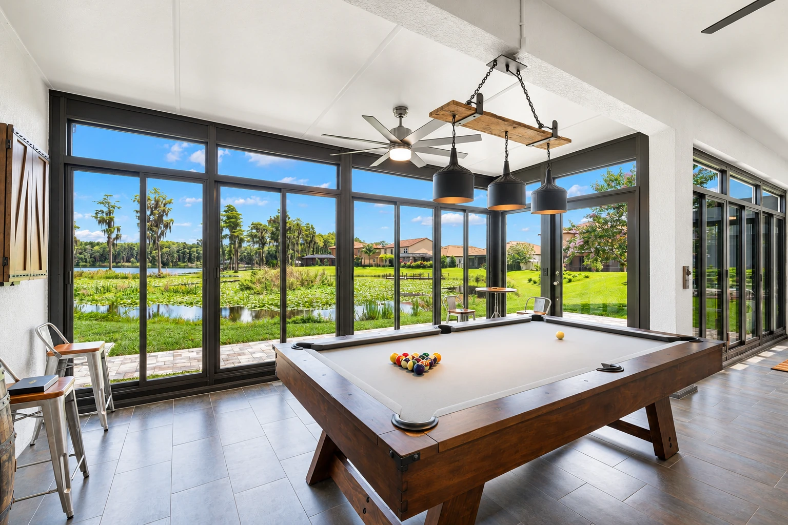 Four-season sunroom with dark-framed floor-to-ceiling windows overlooking a lake, furnished with a pool table and pendant lights.