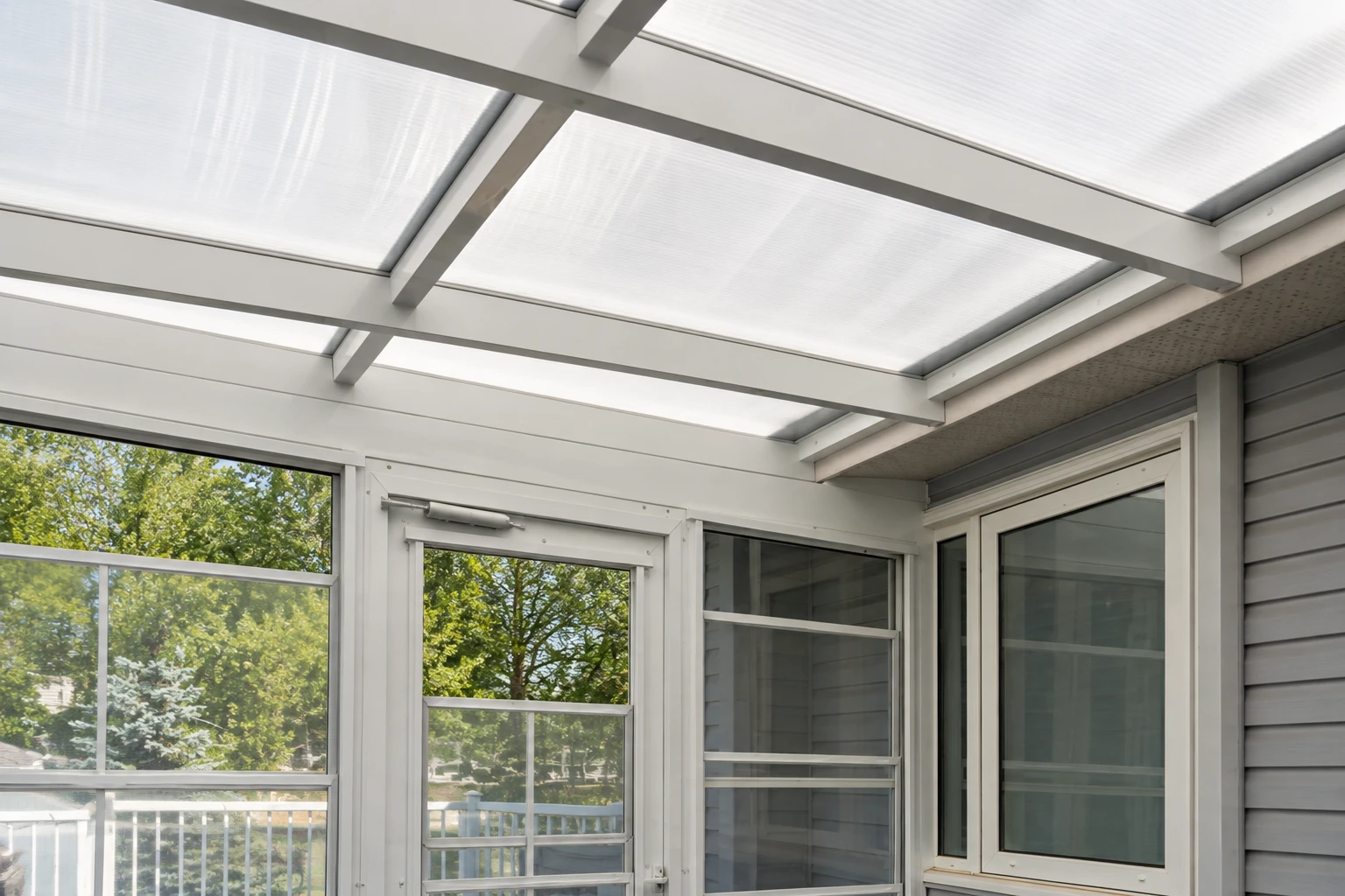 Three-season sunroom showing a translucent paneled roof, screened windows, and white framing beside a gray-sided house.